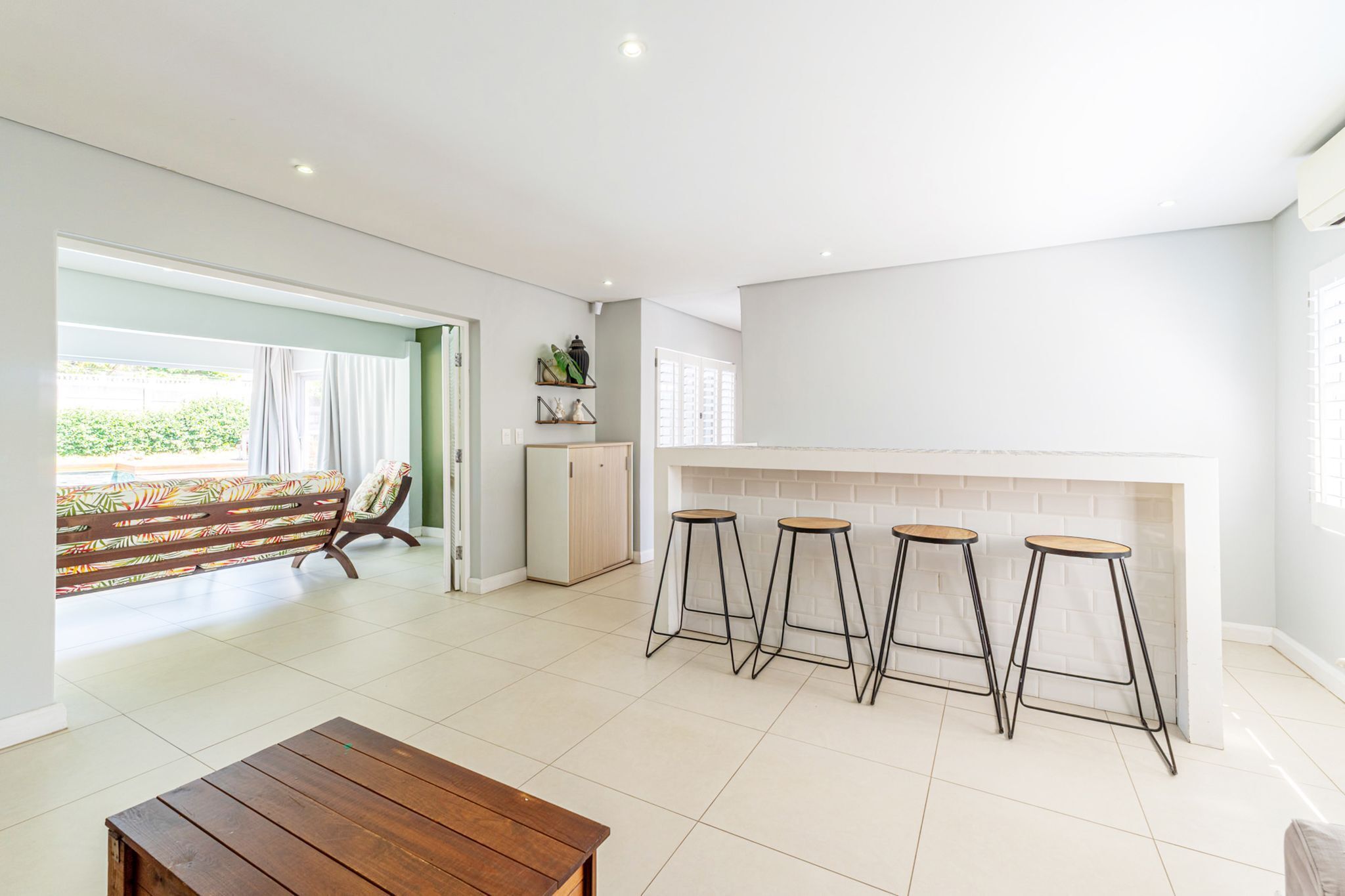 Un vaste espace de vie doté d’une grande fenêtre, d’un banc en bois, d’une cuisine avec un plan de travail blanc et de trois tabourets de bar.