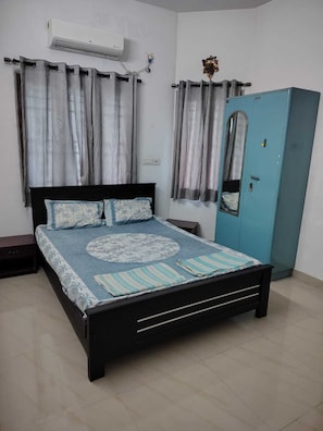 A bedroom with a bed, curtains, an air conditioning unit, a wardrobe, and a nightstand.