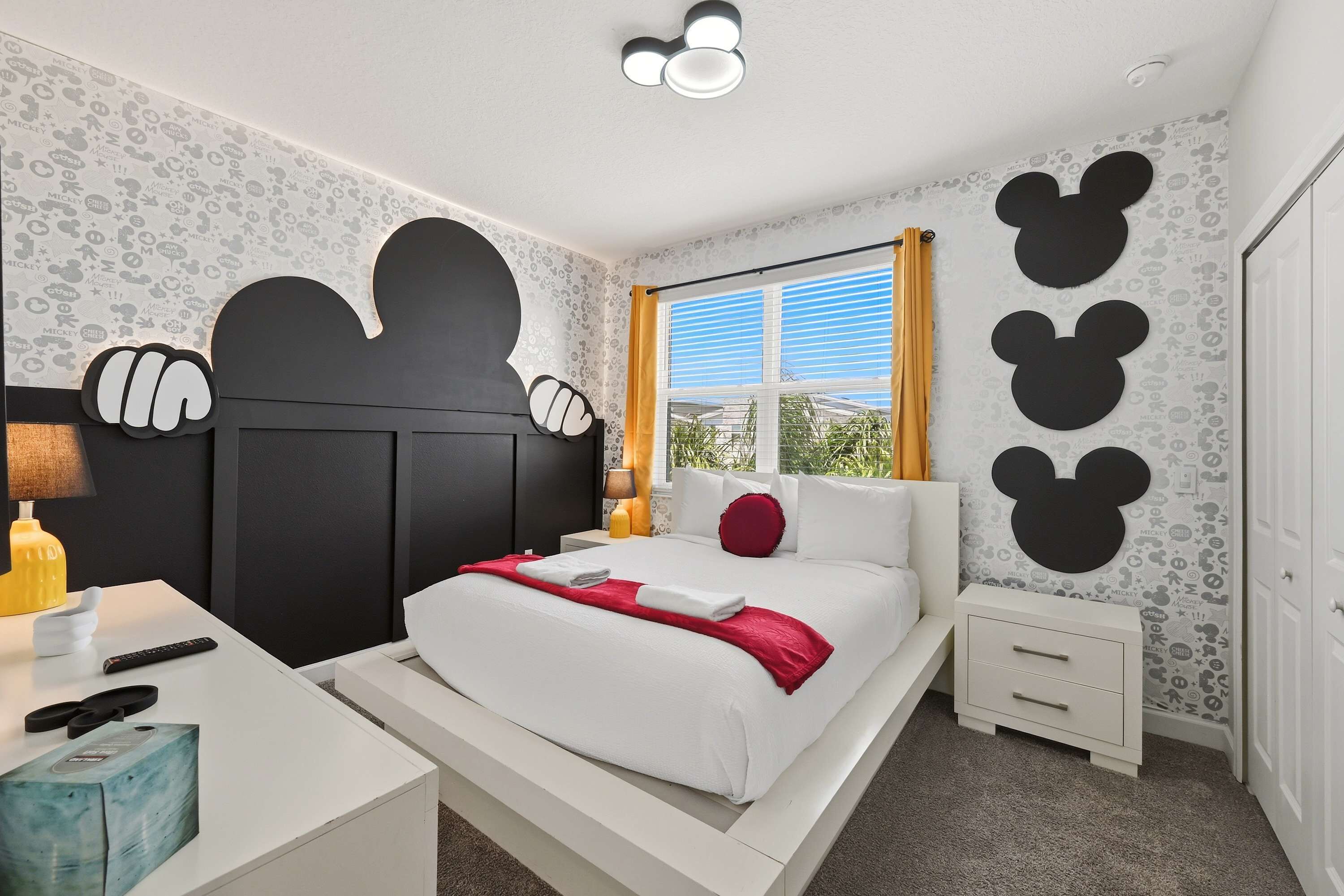 A bedroom with a bed, bedside tables, a nightstand, a window with curtains, and Mickey Mouse wall decorations.