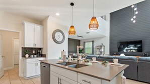 Una cocina moderna con una isla central, iluminación colgante, una zona de comedor con sofá y un televisor montado en una pared oscura.