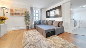 A modern living room with a sectional sofa, a large mirror, a painting of a cityscape, and a kitchen in the background. - 
Cossy two bedroom apartment in Canning Town (London)