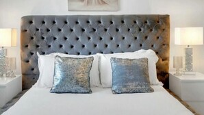 A bedroom with a tufted headboard, a bed with white and blue pillows, and a painting above the bed. - 
Cossy two bedroom apartment in Canning Town (London)