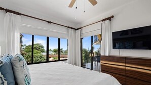 Ein Schlafzimmer mit einem großen Bett, einer hölzernen Kommode, einem an der Wand befestigten Fernseher und großen Fenstern mit Blick auf Gr ünflächen und Gebäude.