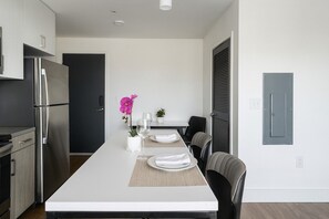A modern kitchen with a stainless steel refrigerator, a dining area with a long table set for four, and a small potted plant on the table.