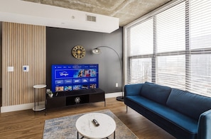 Una sala de estar moderna con un televisor de pantalla plana, un sofá azul, una mesita de centro y un ventanal con persianas.