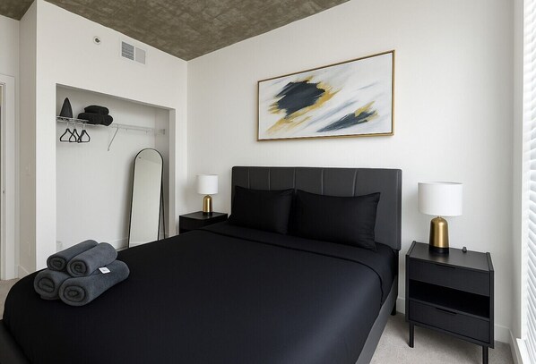 Un dormitorio moderno con una cama grande, dos mesitas de noche, una obra de arte en la pared y un armario con ropa colgada.