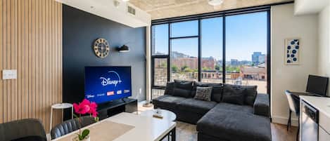 A modern living room with a flat-screen TV, a sofa, a dining table, and a city view through large windows.