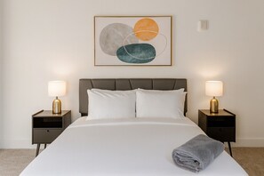 Un lit bien fait, avec du linge de lit blanc, deux tables de chevet avec des lampes et un tableau abstrait accroché au mur.