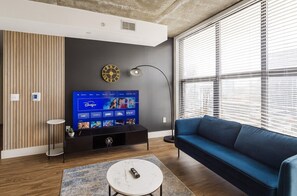 A modern living room with a flat-screen TV, a blue sofa, a coffee table, and a large window with blinds.