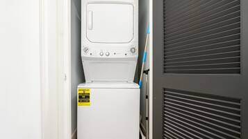 Une petite buanderie blanche, équipée d’un lave-linge et d’un sèche-linge à chargement frontal, avec une étiquette d’avertissement bien visible sur le sèche-linge et une porte entrouverte révélant un mur gris à persiennes.