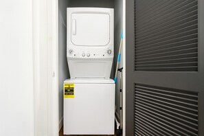 Une petite buanderie blanche, équipée d’un lave-linge et d’un sèche-linge à chargement frontal, avec une étiquette d’avertissement bien visible sur le sèche-linge et une porte entrouverte révélant un mur gris à persiennes.