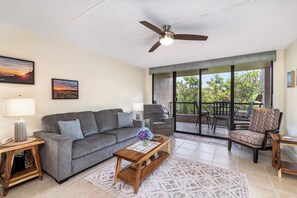 Ein Wohnzimmer mit grauem Sofa, einem Holztisch, einem Deckenventilator und einer großen Glasschiebetür, die auf eine Außenpatio führt.