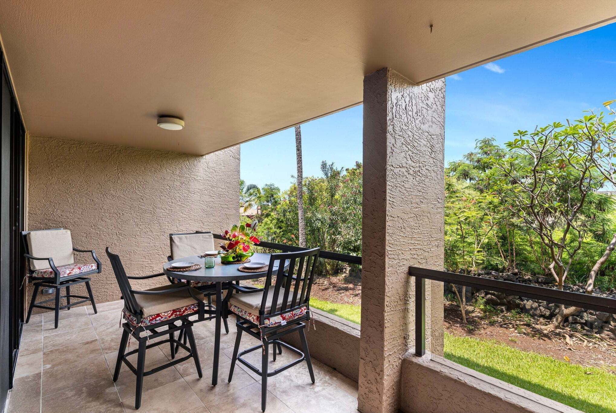 A covered patio with a table and chairs, a flower arrangement, and a view of greenery.