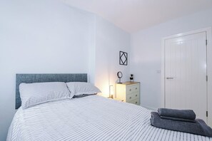 Ein ordentlich bezogenes Bett mit Streifenmuster, ein Nachttisch mit Lampe, eine Kommode und eine Tür.