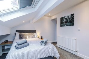 Une chambre avec un puits de lumière, un lit avec des serviettes roulées, une table de chevet, un radiateur et un tableau représentant un paysage urbain.