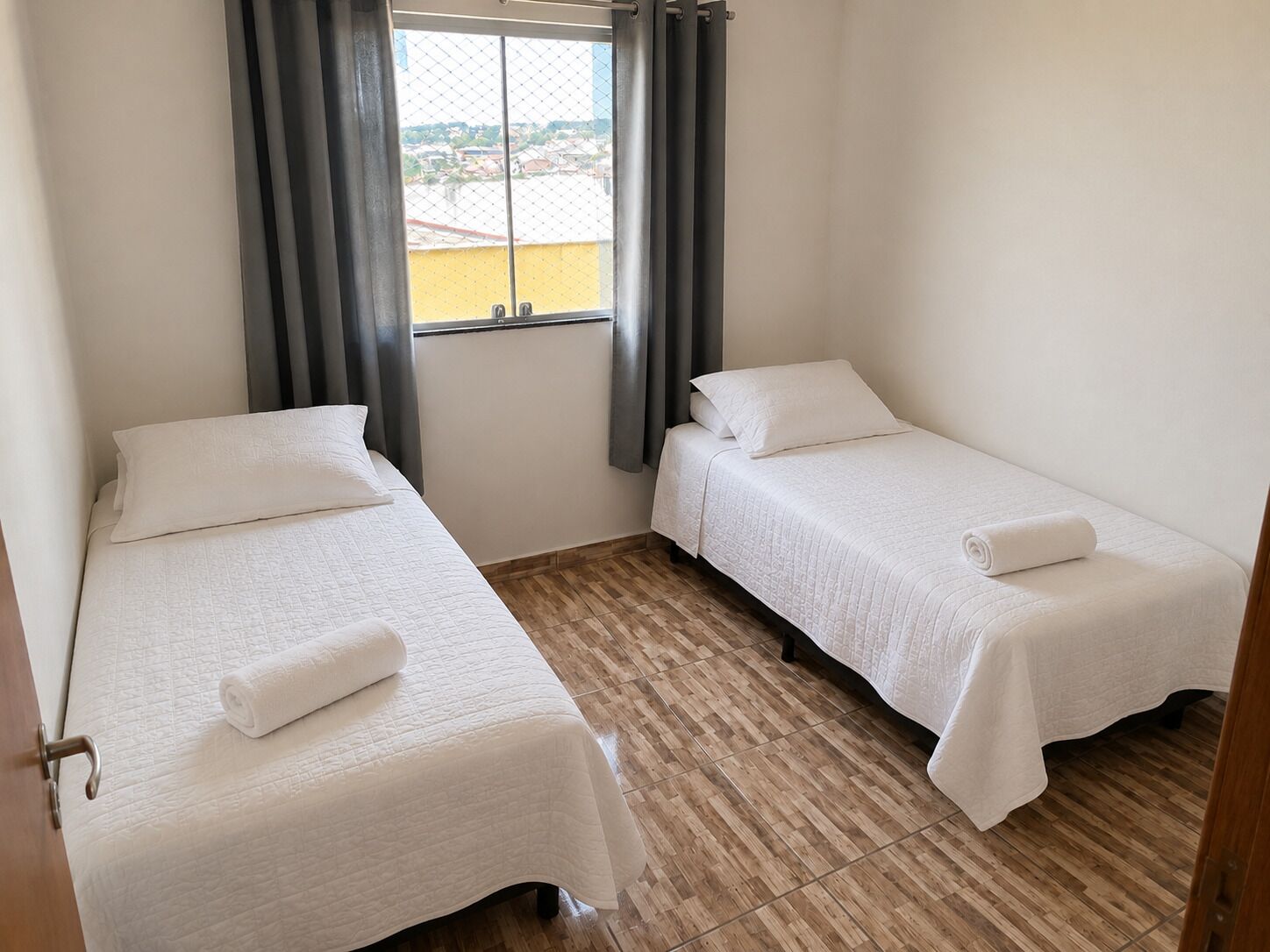 A small, clean room with two single beds, a window with curtains, and a view of buildings outside.