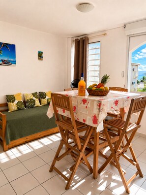A dining area with a table set for four, a sofa with cushions, a window with curtains, and a view of the sea. - Small sandy beach with a pool and ocean view at Anse Mitan (Les Trois-Îlets)