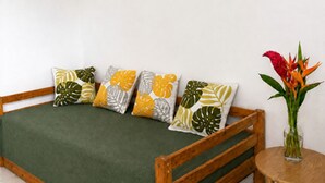 Ein Schlafzimmer mit einem hölzernen Bett, grüner Bettwäsche, tropisch angehauchten Kissen, einem kleinen runden Tisch mit Blumenvase und zwei Wandbildern.
