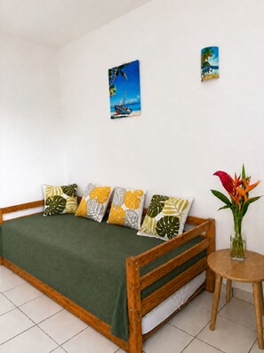 A bedroom with a wooden bed, green bedding, tropical-themed pillows, a small round table with a vase of flowers, and two wall-mounted paintings. - Small sandy beach with a pool and ocean view at Anse Mitan (Les Trois-Îlets)