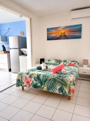 A bedroom with a bed, a painting, a kitchen area, and a bathroom. - Small sandy beach with a pool and ocean view at Anse Mitan (Les Trois-Îlets)