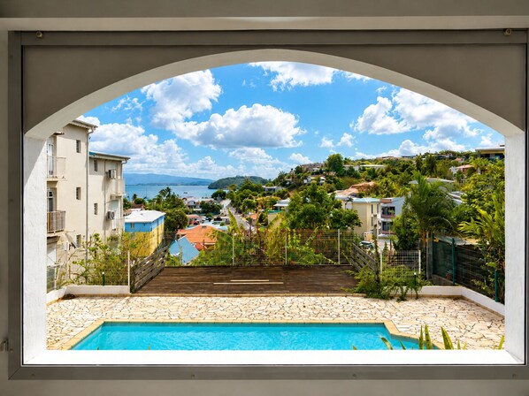 A pool with a stone patio, a view of a coastal town, and a clear sky. - Small sandy beach with a pool and ocean view at Anse Mitan (Les Trois-Îlets)