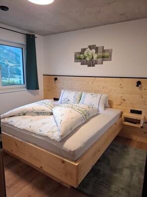 A wooden bed with a headboard, a bedside table, a wall-mounted artwork, and a window with a view of a construction site. - Aussichtsplatz Apartment - Mayrhofen-Hippach Vacation Region (Laimach)