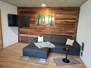 A modern living room with a wooden wall panel, a flat-screen TV, a grey sofa, and a small round table with a pineapple decoration. - Aussichtsplatz Apartment (Laimach)