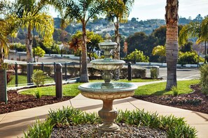 Ein mehrstufiger Brunnen in einem angelegten Garten mit Palmen und Blick auf eine hügelige Landschaft.