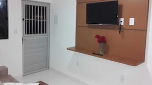 A room with a closed metal gate, a mounted television on a wooden shelf, and a small vase with red flowers. - FLATS CABANOS (Maragogi)