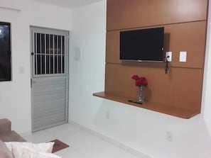 Um quarto com um portão de metal fechado, uma televisão fixada numa prateleira de madeira e um pequeno vaso com flores vermelhas.