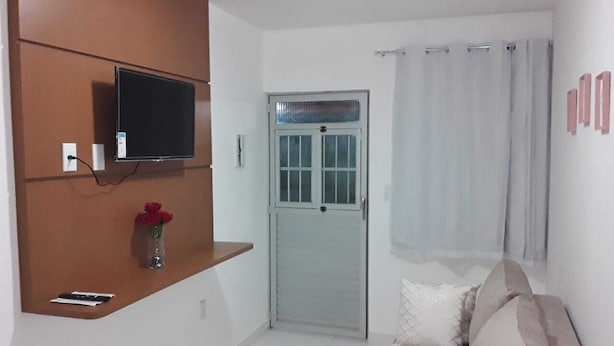 A living room with a flat-screen TV mounted on a wooden panel, a door with a glass panel, a sofa with cushions, and a small vase with flowers on a shelf. - FLATS CABANOS (Maragogi)