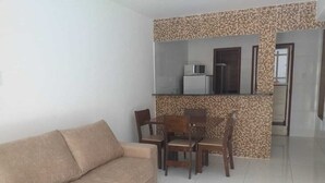 A living room with a beige sofa, wooden dining table, and chairs. A kitchen area is visible in the background with a refrigerator and microwave. - FLATS CABANOS (Maragogi)