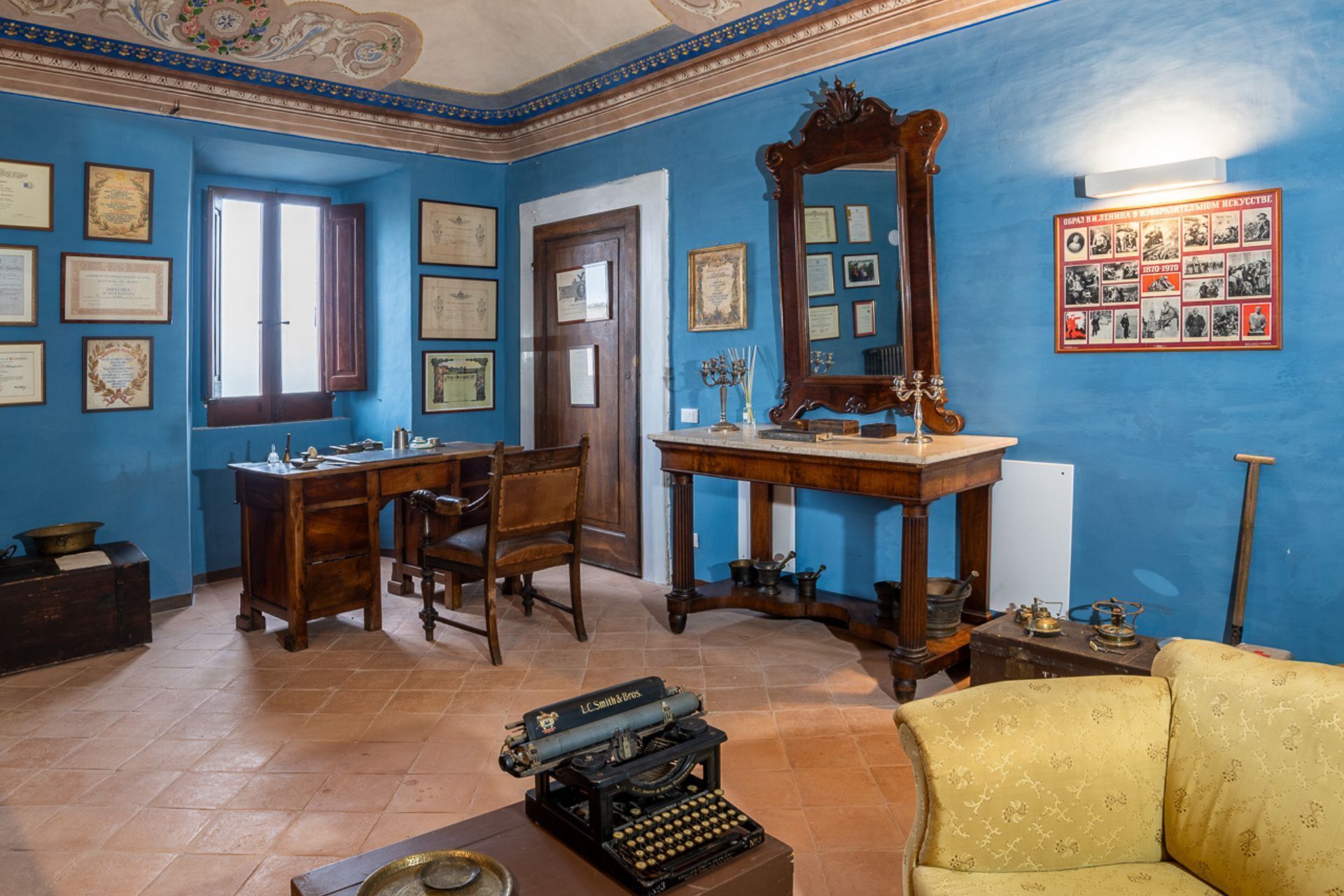 A room with a blue wall, wooden desk, a typewriter, a mirror, and framed certificates.