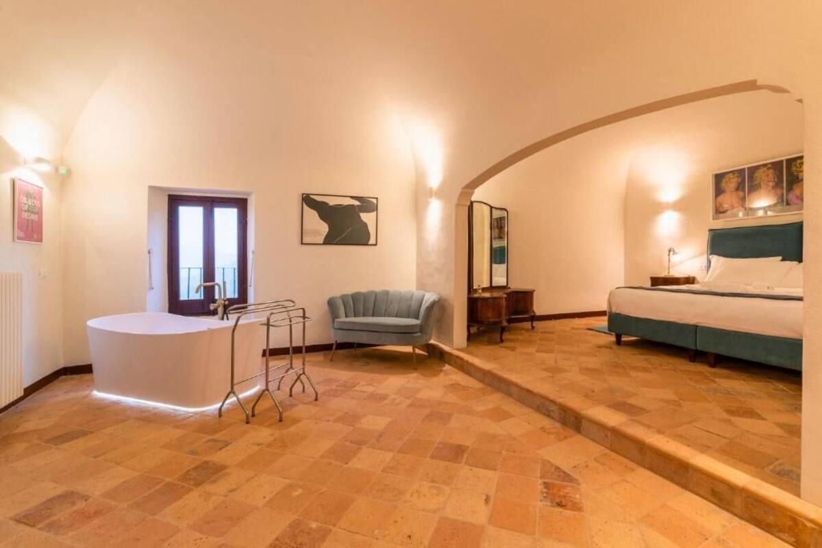 A hotel room with a curved ceiling, a bed, a chair, a small table, and a bathroom with a sink and towel rack.