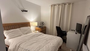 A neatly made bed with white linens, a bedside lamp, a small desk with a computer and a fan, and a television on a stand. - Sleeps4-familyfriendly-kingbeds-2bath (London)