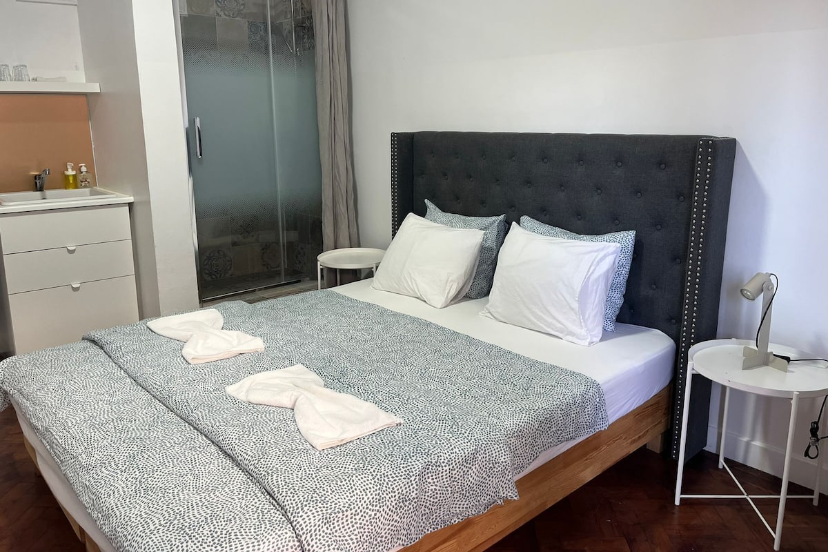 Habitación de hotel con cama, mesita de noche, espejo, armario y zona de ducha.
