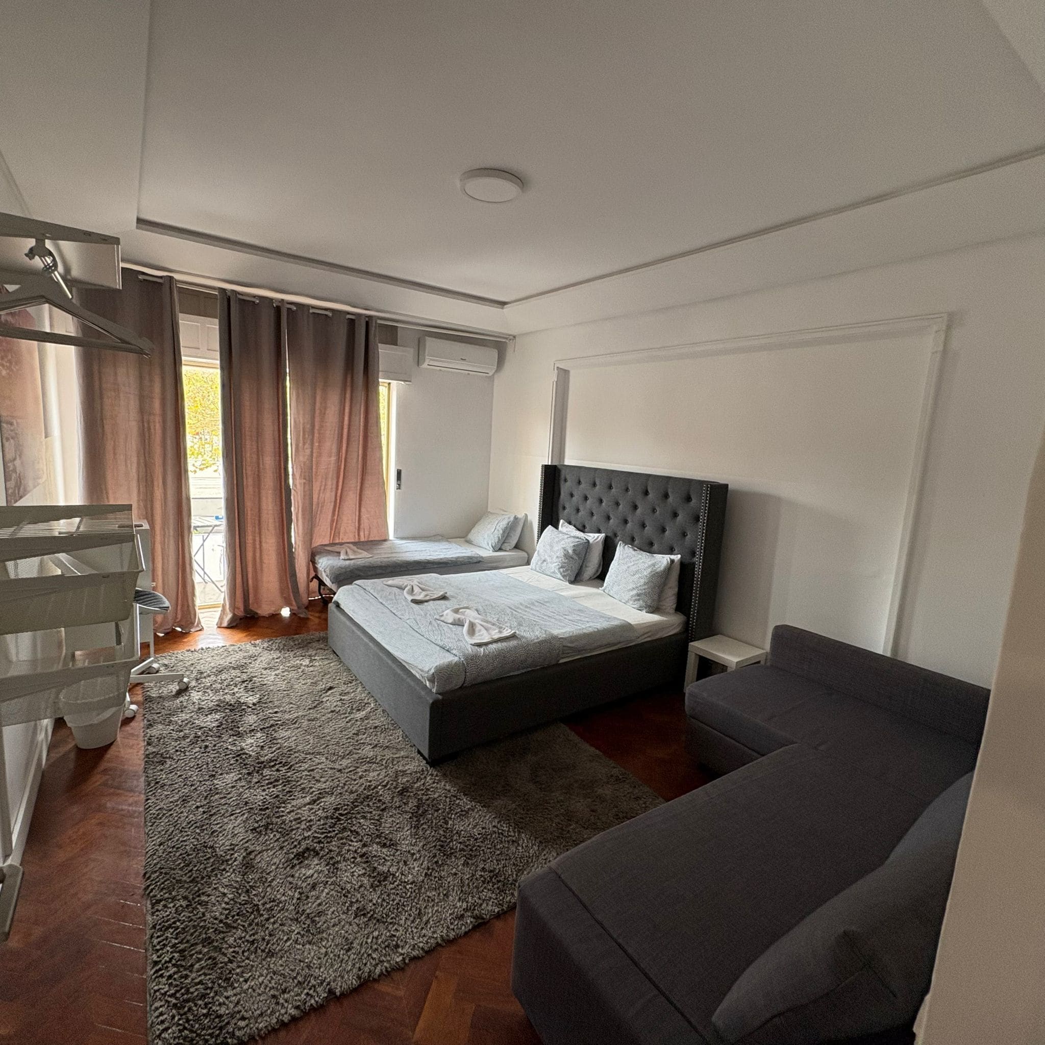 A bedroom with a bed, two bedside tables, a chair, a sofa, a window with curtains, and an air conditioning unit.