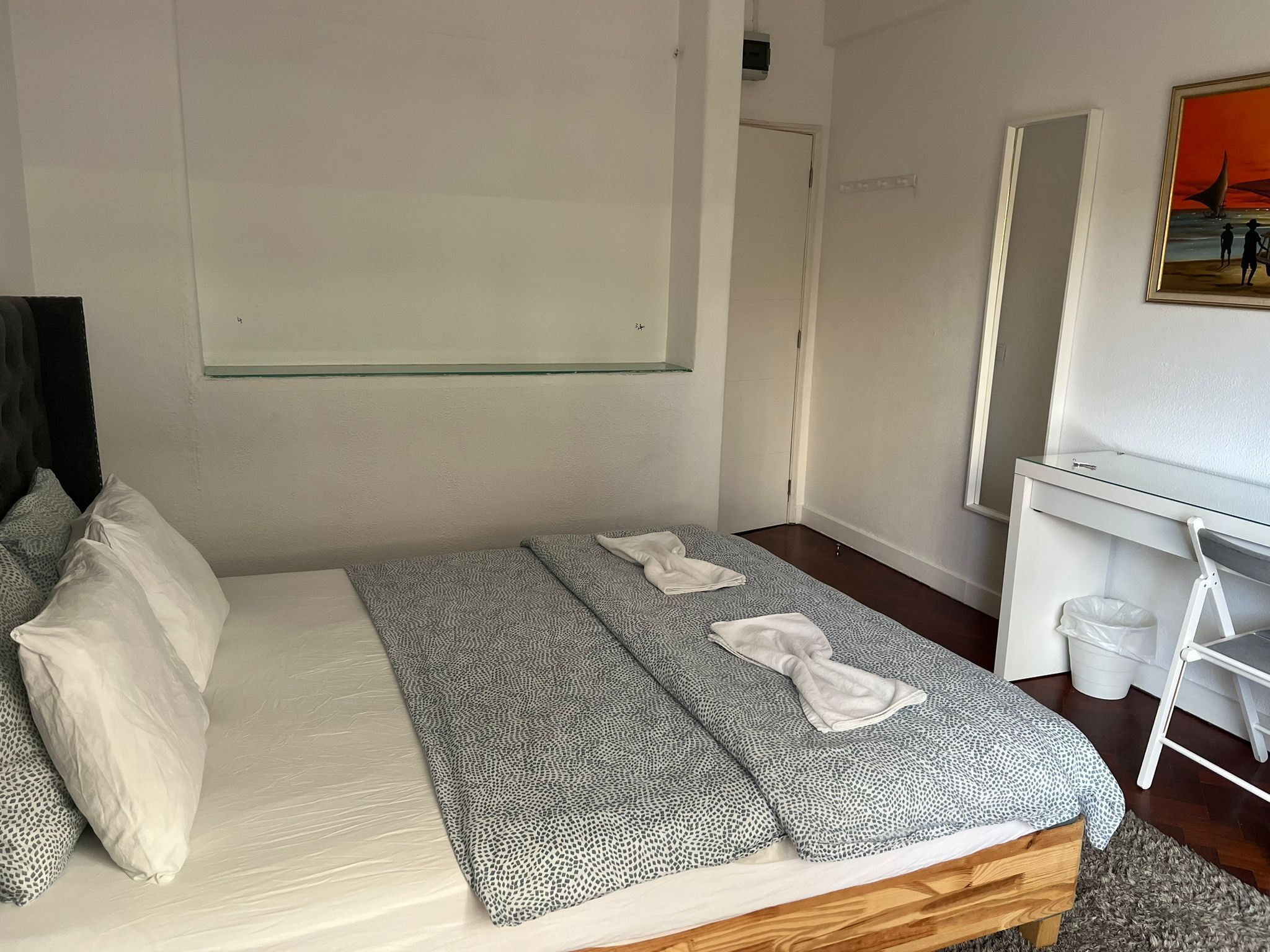 A neatly made bed with patterned bedding, a bedside table with a mirror, and a wall-mounted shelf.