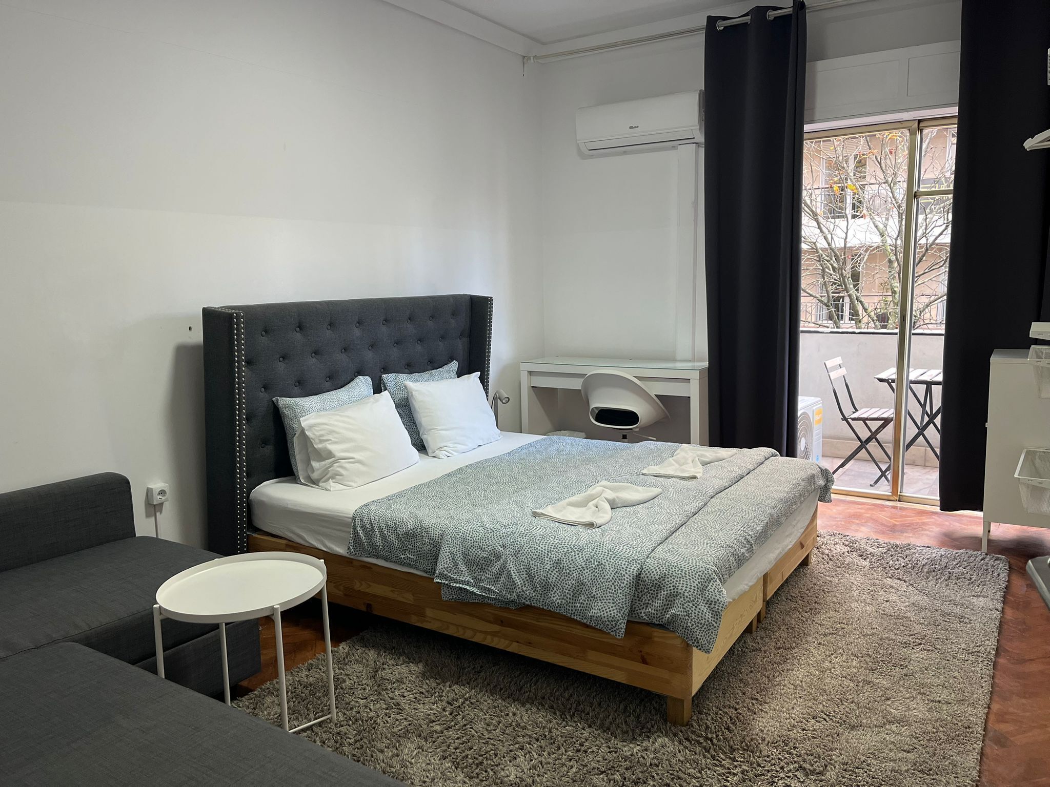 A bedroom with a bed, a small table, a sofa, a desk, and a window with a view.