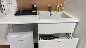 Angolo cottura con piano di lavoro bianco, lavello, piccolo frigorifero e cassetto con stoviglie.