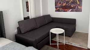 Una camera d'albergo moderna con un divano grigio, un piccolo tavolino bianco, un letto con biancheria a motivi e un quadro raffigurante un panorama urbano.