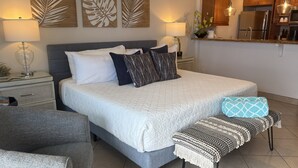 Un dormitorio con cama, mesitas de noche, un banco y un rincón cocina.