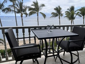 Varanda com mesa e cadeiras, vista para uma praia e oceano com palmeiras.