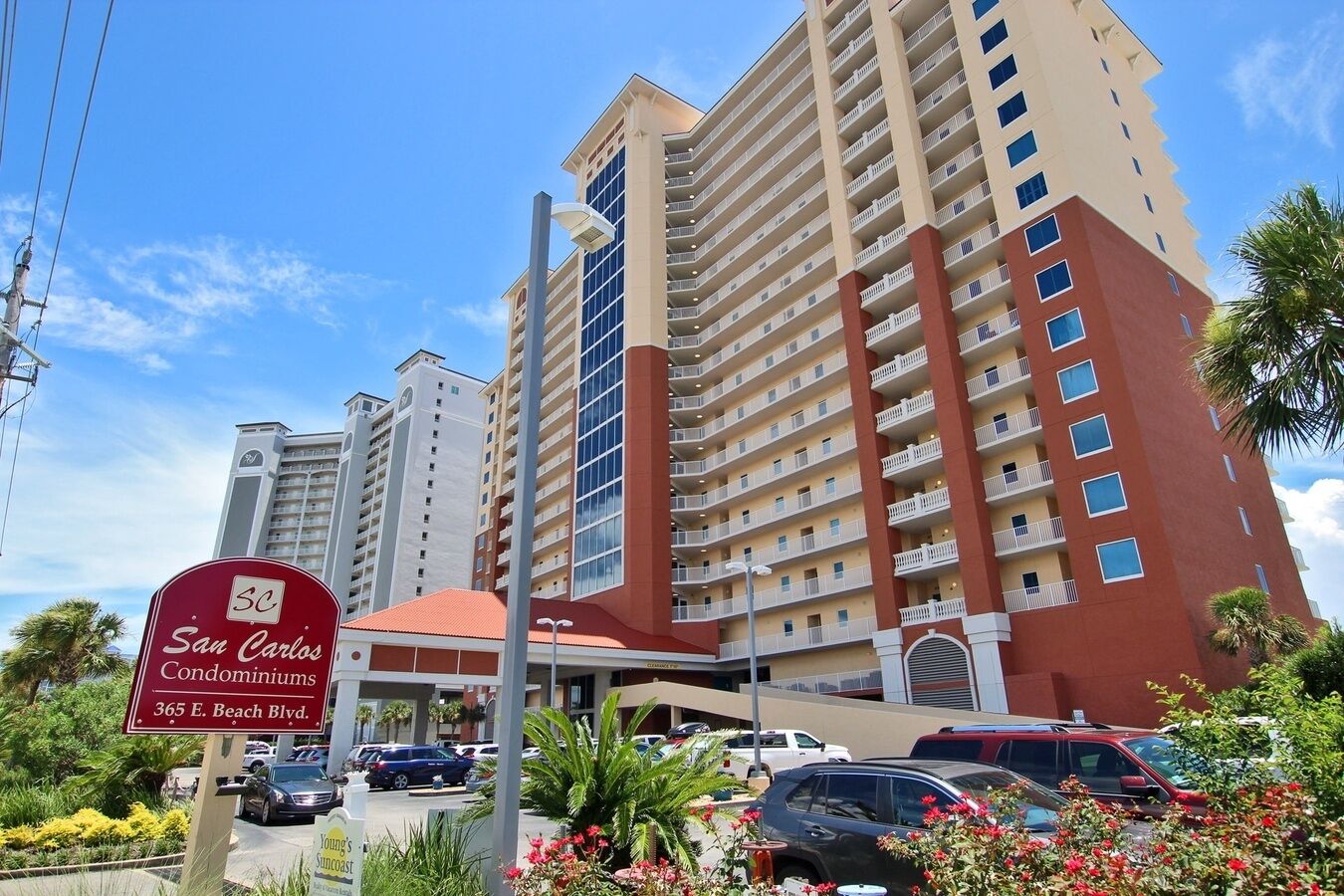 Un hôtel à plusieurs étages, doté d’une façade rouge et beige, d’une enseigne indiquant « San Carlos Condominiums, 365 E. Beach Blvd. », et d’un parking où sont stationnées plusieurs voitures.