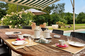 Un área de comedor al aire libre con una mesa de madera dispuesta para un servicio de té, rodeada de sillas de mimbre y un gran paraguas.