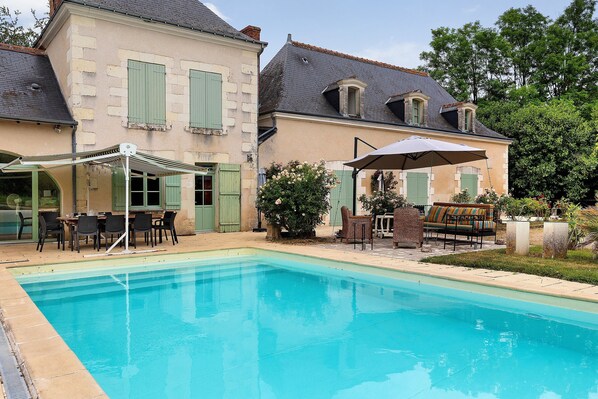 A swimming pool with a patio area, outdoor furniture, and a building with shutters.