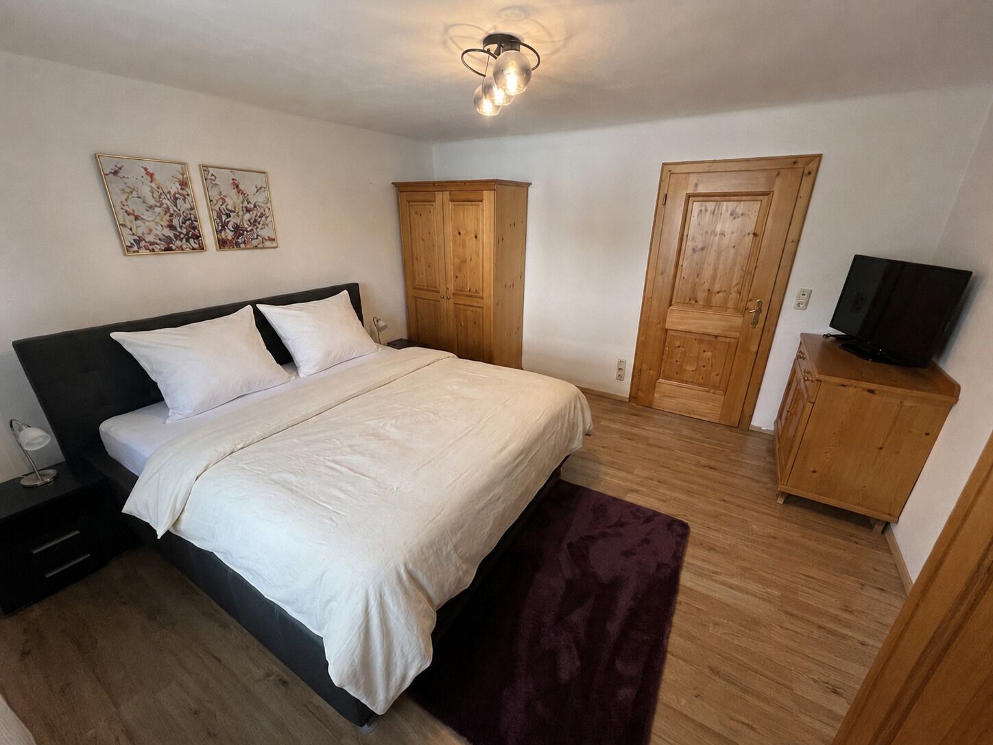A bedroom with a bed, wooden wardrobe, a television, and two wall-mounted paintings.
