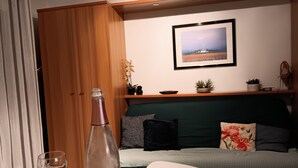 Une pièce comprenant un meuble en bois, une bouteille de champagne, un verre, une petite table, un canapé avec des coussins, un tableau encadré au mur et une étagère avec des plantes.