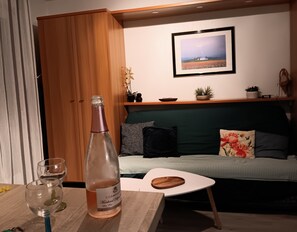 Ein Zimmer mit einem Holzkasten, einer Champagnerflasche, einem Glas, einem kleinen Tisch, einer Couch mit Kissen, einem gerahmten Bild an der Wand und einem Regal mit Pflanzen.