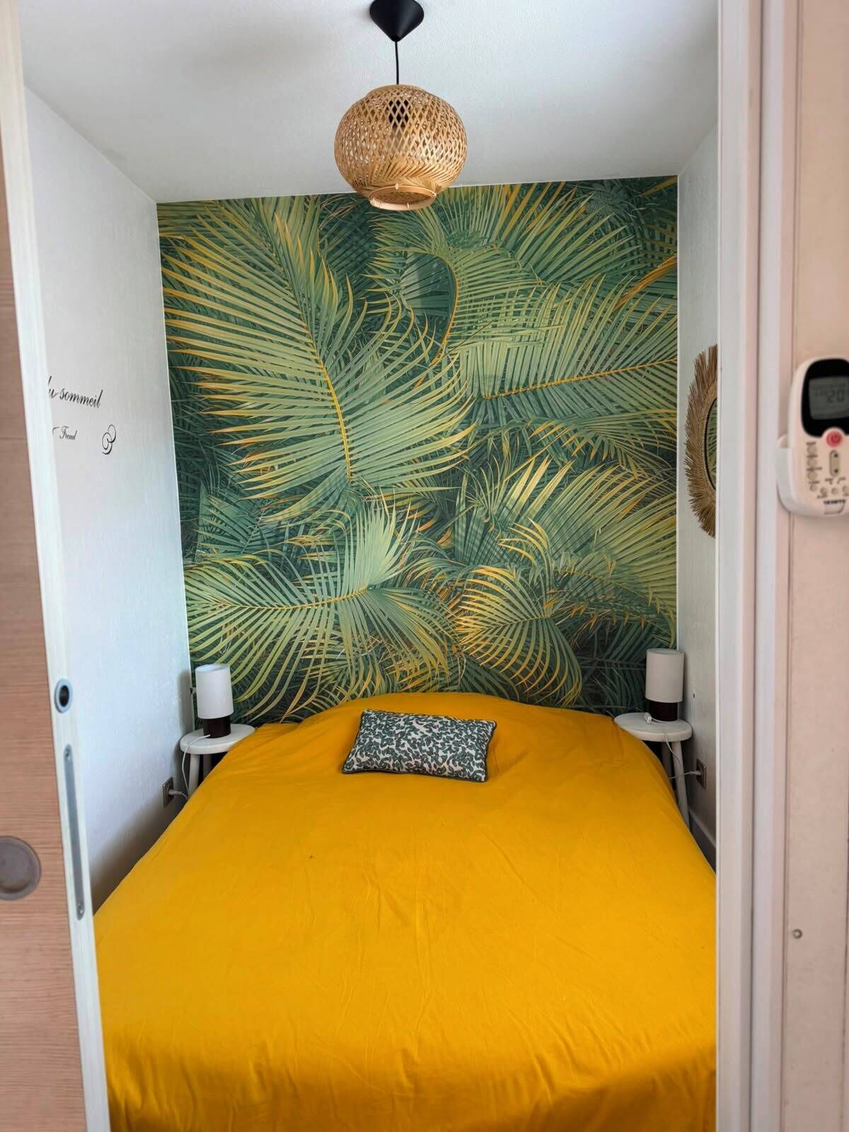 Ein Schlafzimmer mit gelber Bettdecke, einer Wandweberei mit Palmenblättern, einer hängenden Rattan-Leuchte, Nachttischlampen und einem Thermostat.